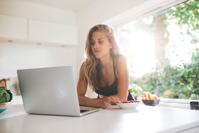 Young woman leaning on kitchen counter and looking at laptop
