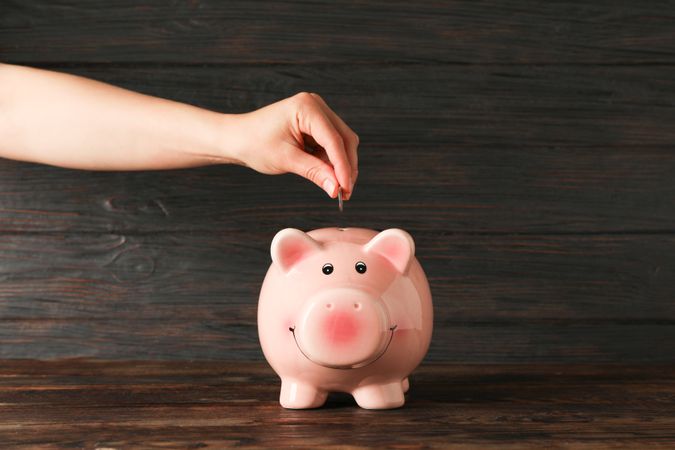 Woman's hand puts a coin in the happy piggy bank on wood table against wooden background, space for text. Finance, saving money