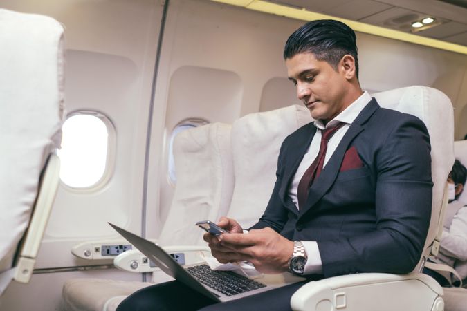 Businessman in suit checking phone in airplane with laptop