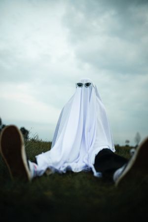 Person wearing ghostly textile with sunglasses sitting on green grass