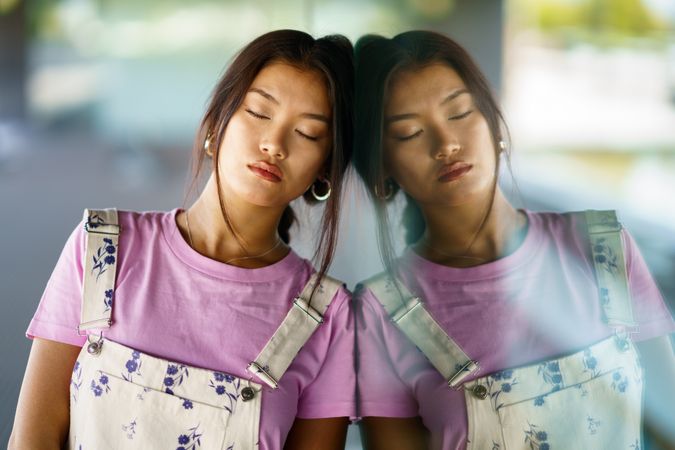 Chinese female in floral overalls and pink t-shirt leaning against glass on building outside