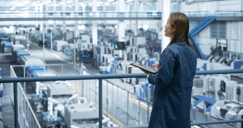 Japanese Factory Manager Working on a Tablet at a Futuristic Industrial Manufacture with Autonomous AI Robots. Specialist Monitoring All the Data on a Device, Checking Reports and Planning Research