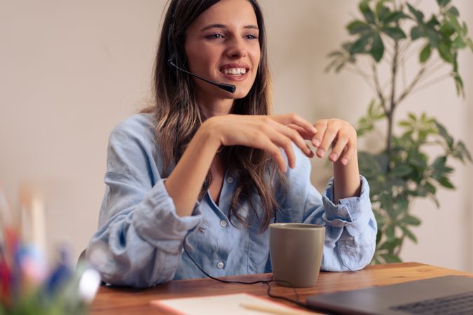 Enthusiastic young female professional enjoying virtual team meeting and coffee break while working remotely in modern home office