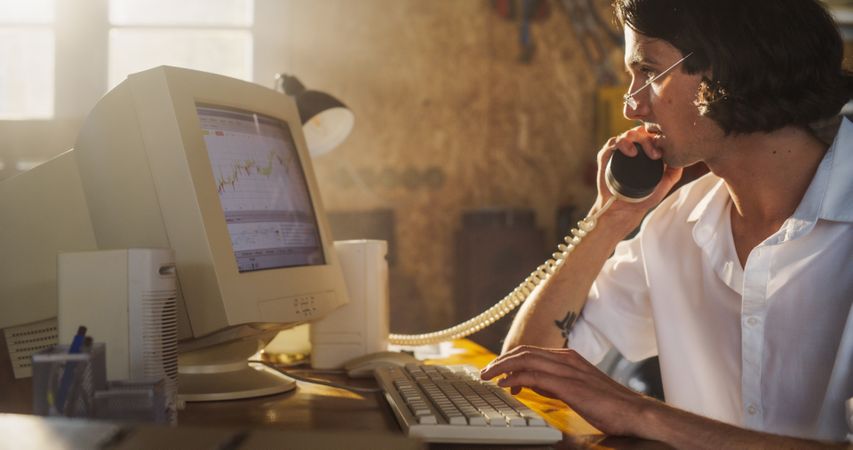 Male Trader Talking On Landline Phone And Using Old Desktop Computer In Retro Garage.