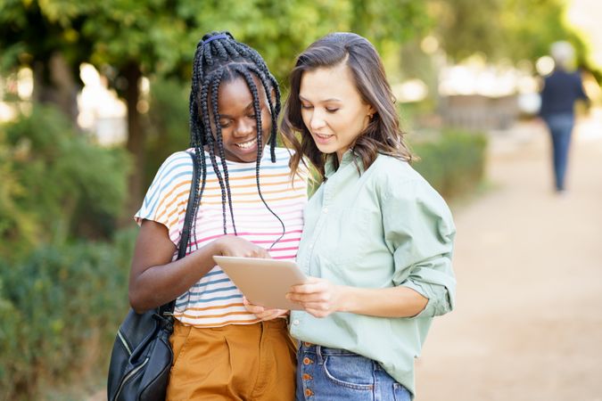 Women friends pointing at something on tablet in casual clothing outside