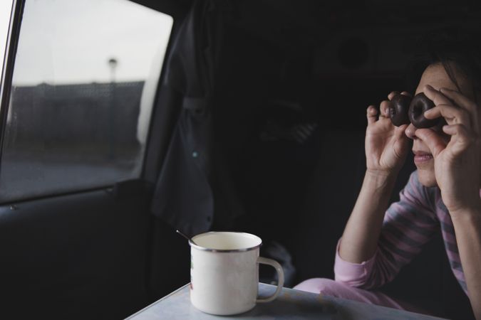 Woman with biscuits near eyes sitting in car with cup