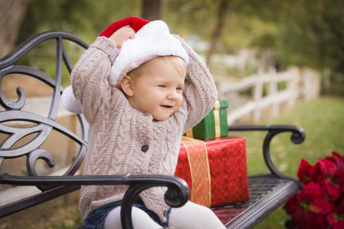 Young Child Wearing Santa Hat Sitting with Christmas Gifts Outside.