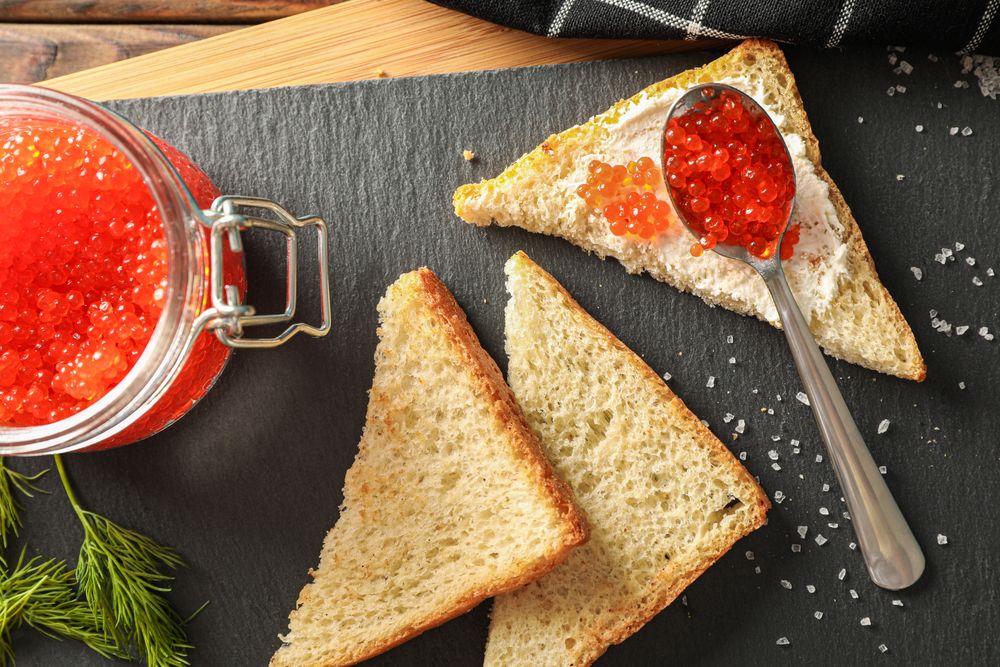 Composition with caviar in glass jar, bread, spoon, towel and cutting board, top view