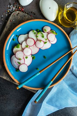 Top view of sliced radishes served with chopsticks on blue plate with side of oil