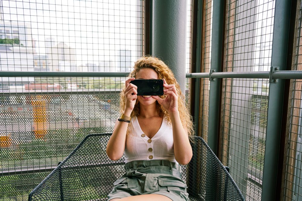 Pleasant woman taking photo with smartphone sitting in metal chair