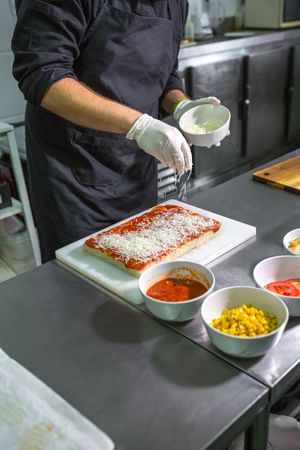Unrecognizable male chef sprinkling grated cheese over tomato sauced focaccia in restaurant kitchen