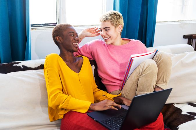 Female couple laughing together on sofa with laptop and notebook