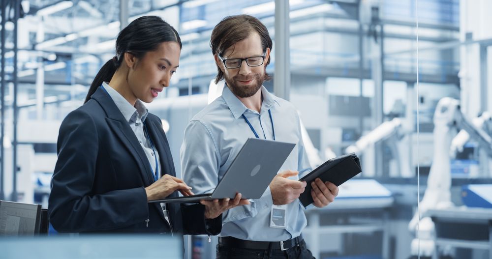 Industrial Engineers Working at Technology Factory. Man and Asian Female Using Laptop and Tablet Computers and Having a Conversation. Footage with a Portrait of a Couple of Employees
