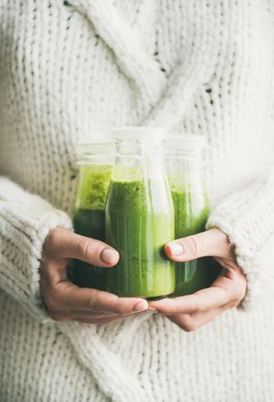Woman holding three bottles of green smoothie wearing cozy sweater
