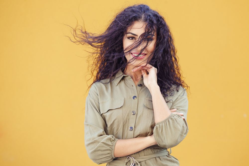 Female in army green jumpsuit smiling in front of mustard wall with hair blowing