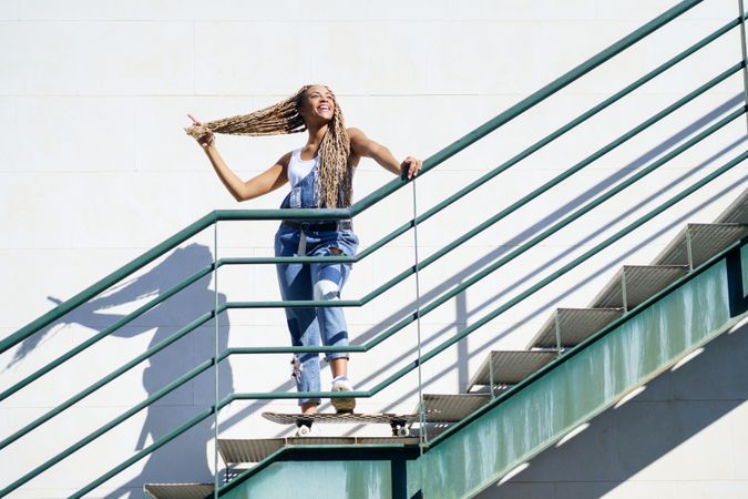 Happy female in denim overalls standing on skateboard on stairs playing with her hair
