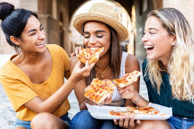 Three cheerful multi-ethnic women eating pizza in the street