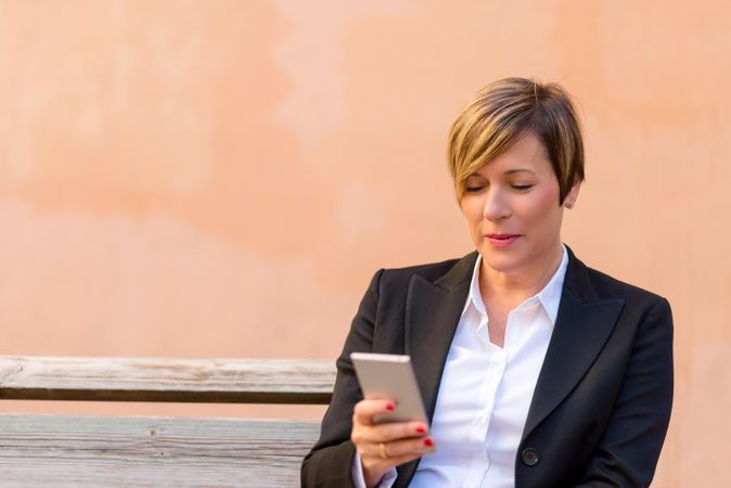 Woman in blazer sitting on bench checking phone in front of peach wall