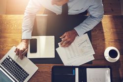 Overhead shot of man in business attire working on his laptop with documents and coffee on desk