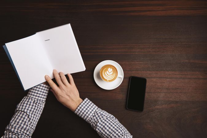 Arms in plaid shirt holding notebook with cellphone and coffee on table