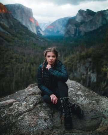 Young woman in dark bubble jacket sitting on rock formation near lake
