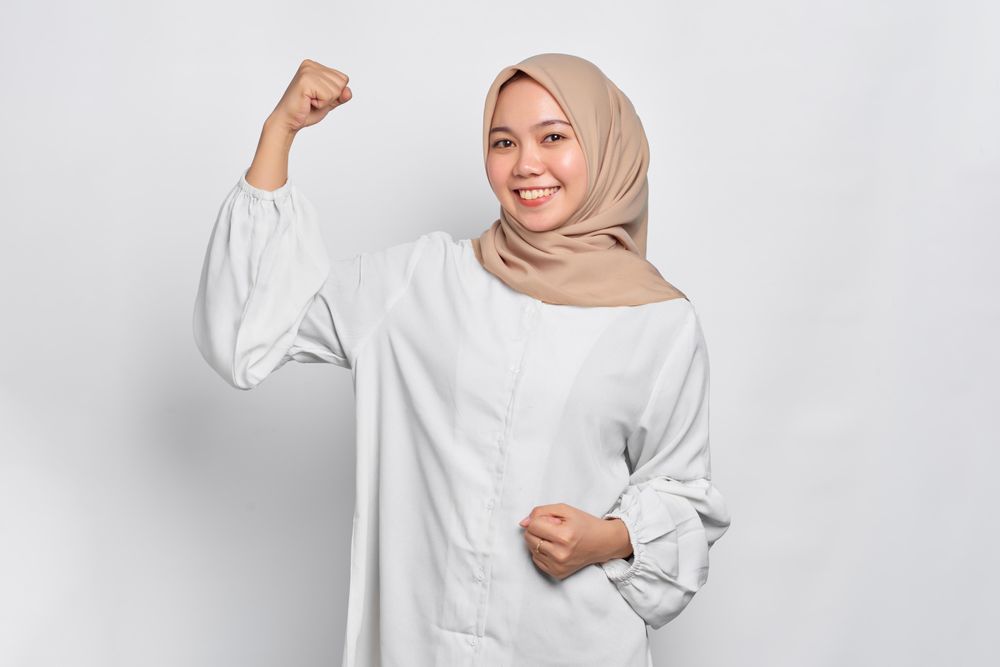 Muslim woman in headscarf and light blouse and smiling and flexing her biceps