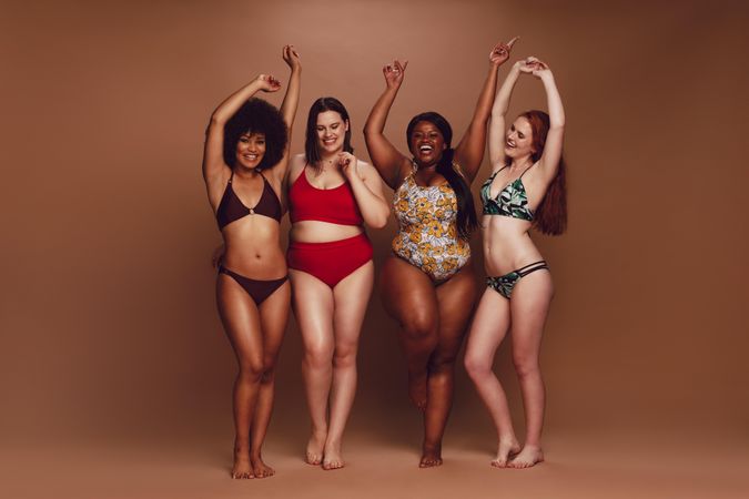 Full length of slim and curvy women in bikinis dancing together over brown background