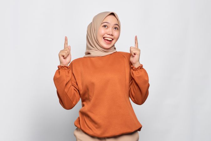 Happy Muslim woman in headscarf and orange sweater pointing upwards with good idea