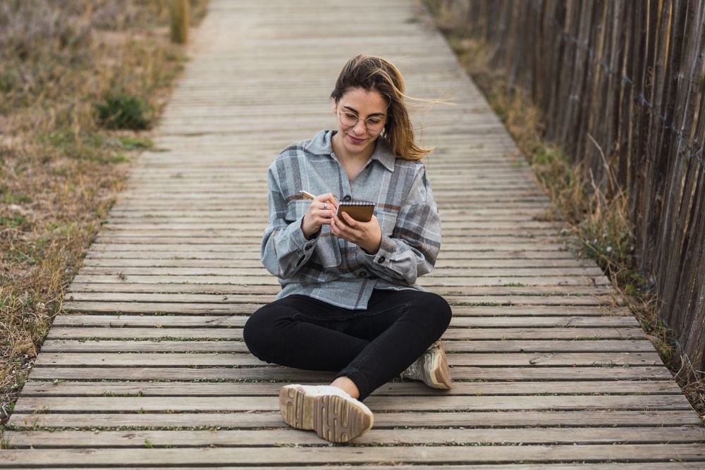 Dreamy woman sitting on boardwalk and writing in notebook in nature