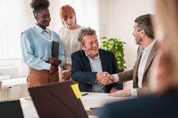 Multi-ethnic business professionals shaking hands in agreement, with attentive colleagues in the background