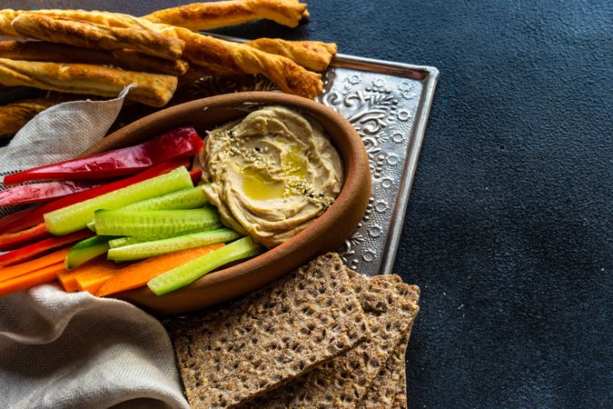 Creamy hummus dish served with chopped veggies, rye crackers and bread sticks on trey