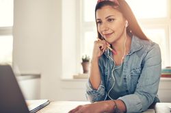 Contemplative woman reading on laptop in home office with earbuds and pencil