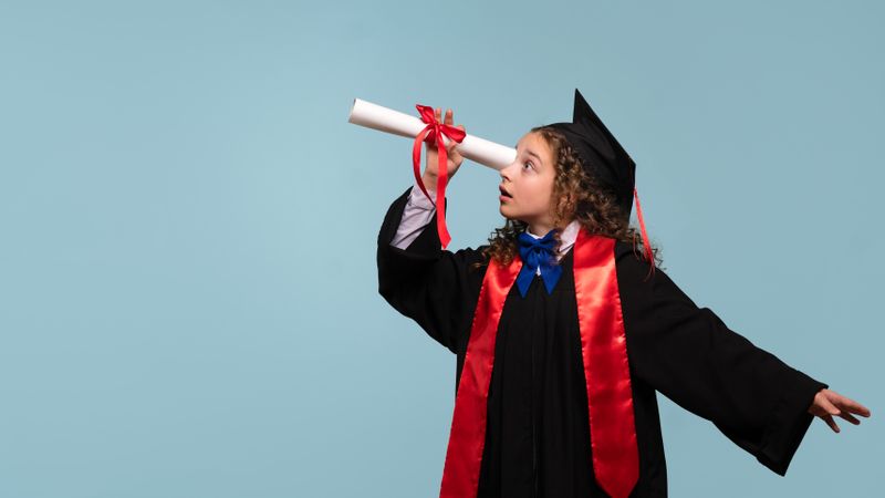 Young girl in graduation attire looking through diploma like telescope.