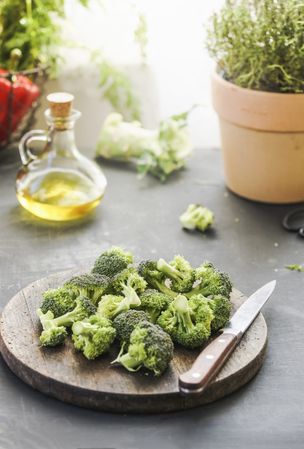Broccoli at wooden cutting board with kitchen knife at grey kitchen table with herbs, oil and ingredients at background with natural lights