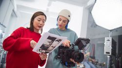 In A Modern Studio, An Asian Woman In A Red Sweater And A Woman With Blue Hair, Both Young, Collaborate Over A Camera And Storyboard, Focusing On Creative Film Production And Advertising.