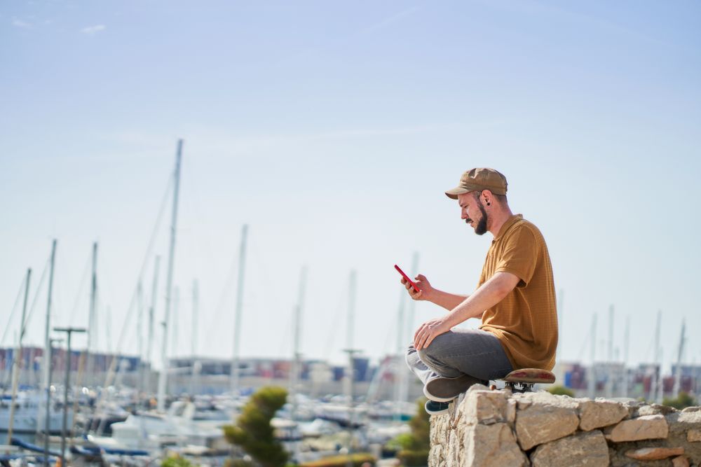 Man in cap relaxed sitting on skateboard using mobile phone