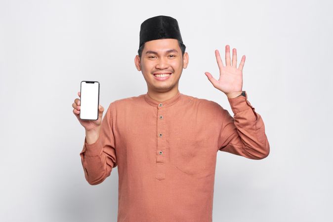 Happy Muslim man in kufi hat with mobile phone waving with other hand