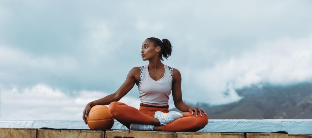 Woman in fitness wear sitting with orange ball outdoors