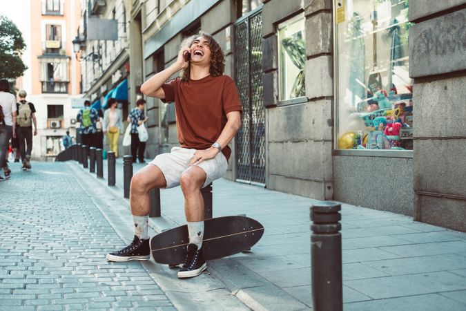 Amused man with longboard talking on phone while resting on street