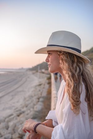 Charming woman in hat leaning on fence and looking at sea