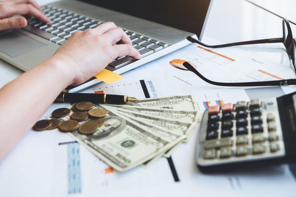 Person’s hands typing on laptop at desk with money, paper and calculator