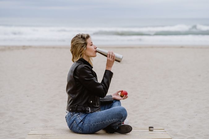 Content woman drinking and eating apple sitting with crossed legs on beach