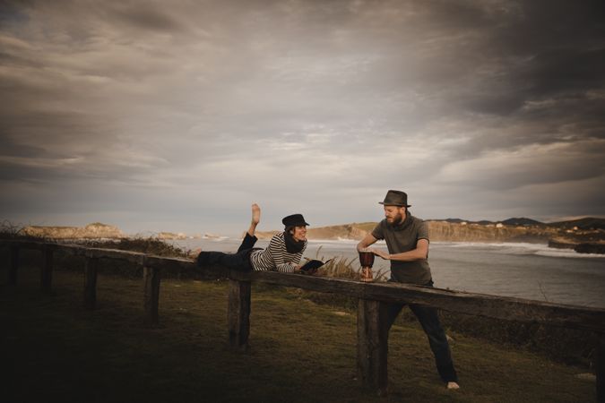 Man plying on drum near woman with notebook on bench on shore