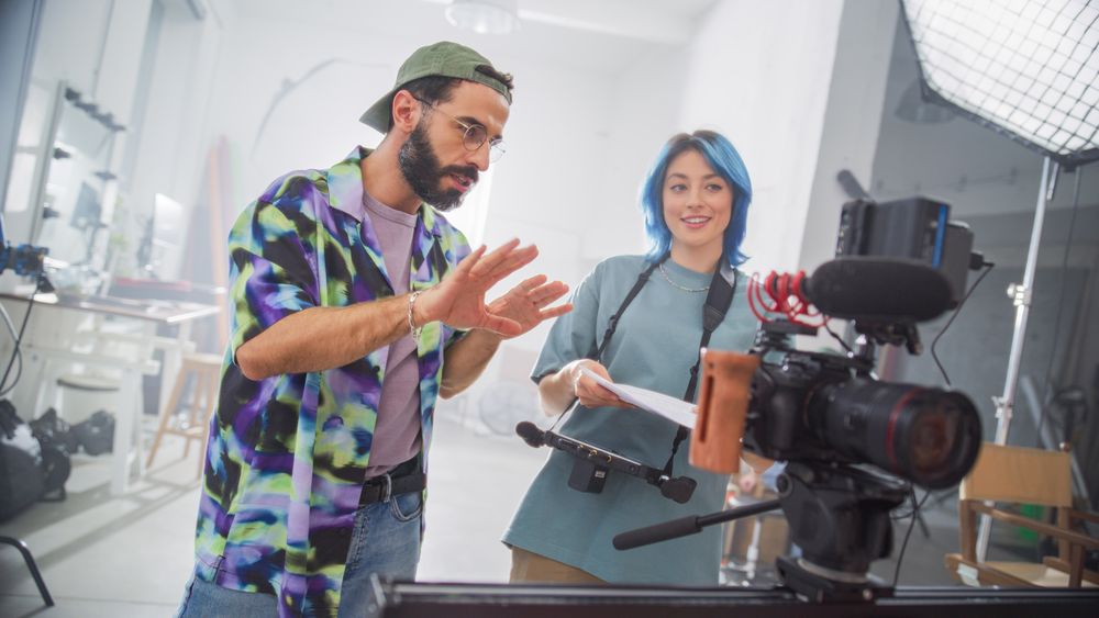 Young Male Director With Beard And Caucasian Female With Blue Hair Discuss Over A Script In A Well-equipped Film Studio, Surrounded By Professional Camera And Lighting Equipment.