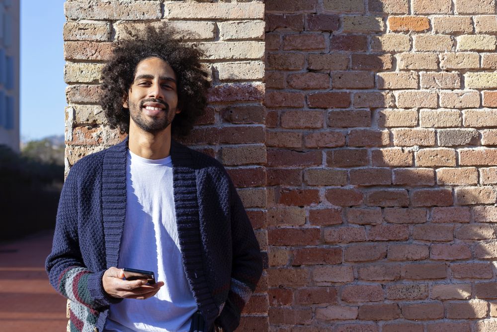 Smiling man holding his smartphone while leaning on a brick wall outdoors on sunny day