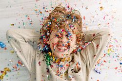 Happy boy covered with colorful confetti lying on floor