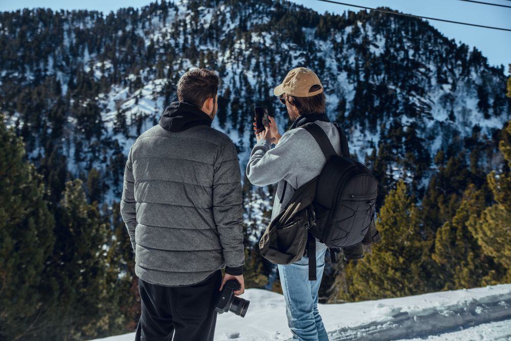 Men with smartphone and camera on hill between trees in winter