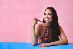 Hispanic woman with long brown hair holding large make up brush and looking away from camera