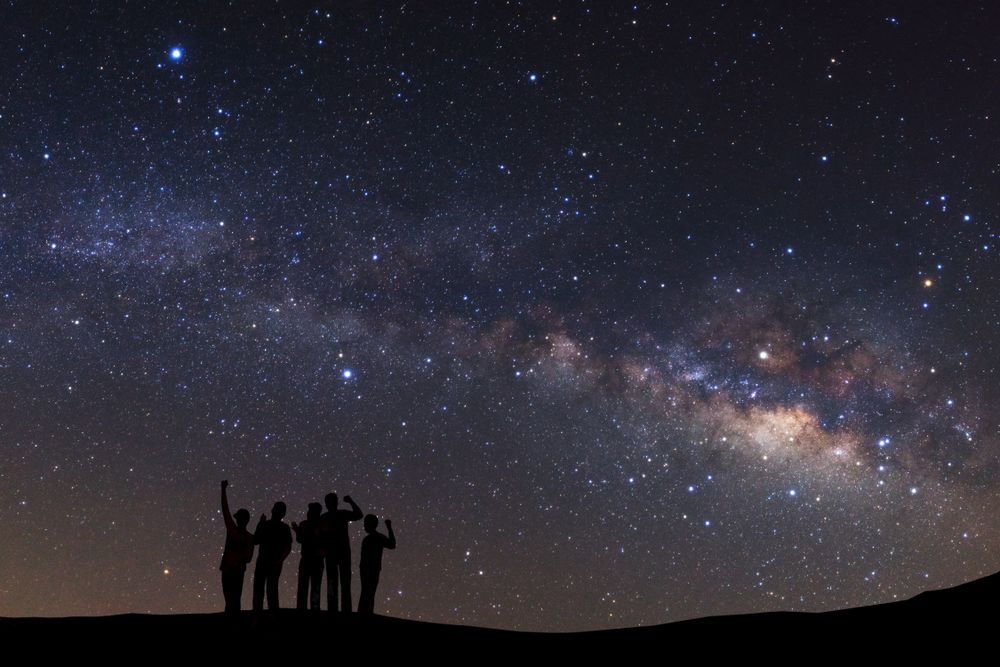 Night sky with stars and silhouette of a standing sporty man with raised up arms on high mountain.