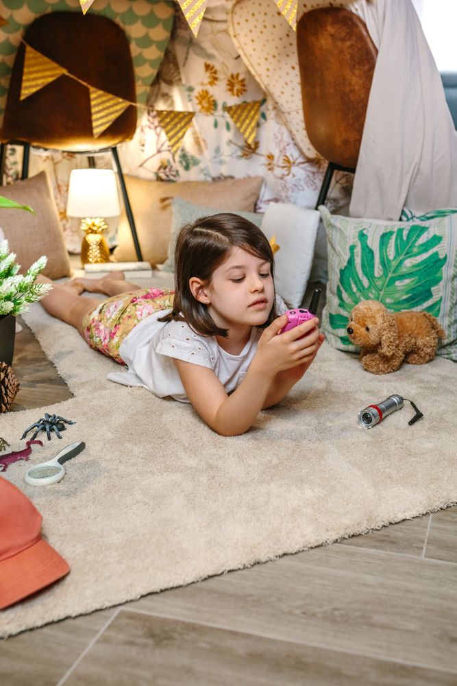 Little girl using walkie-talkie lying over rug surrounded by cushions and plants in teepee at home